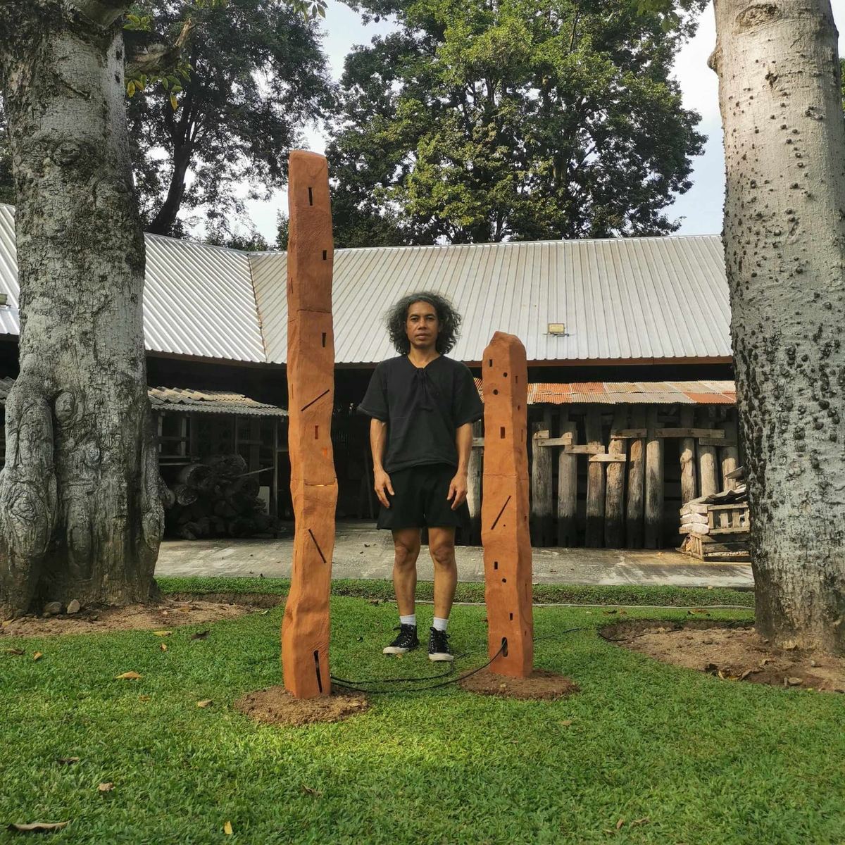 Artist Samran standing between two carved wooden sculptures in the garden, part of the SEN exhibition