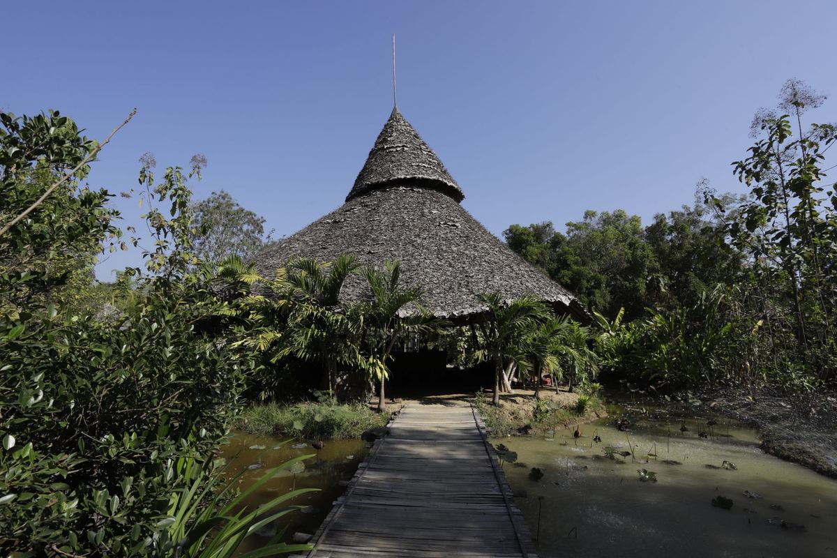 A tall thatched roof at the end of a wooden walkway over the pond
