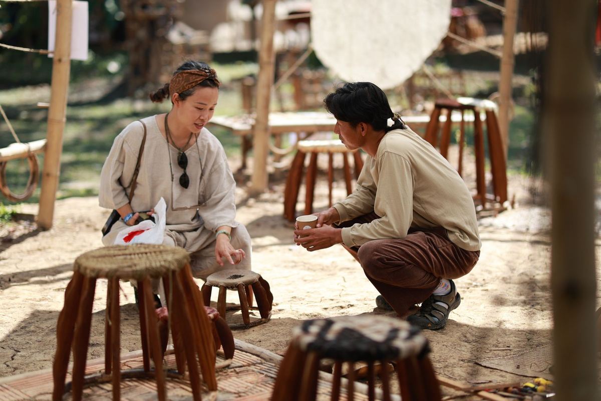 A maker and a visitor talking among handmade wooden stools at a stall